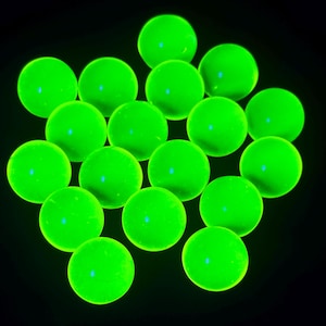 Peut inclure: Une collection de dix-sept billes en verre lisses et sphériques qui brillent d'une lumière vert néon intense et vibrante sur un fond noir uni. Chaque bille présente une forme circulaire lumineuse uniforme avec une surface réfléchissante.