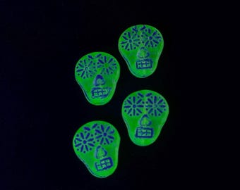 May include: Four neon green skull-shaped objects with blue floral and geometric designs. The skulls have eye and nose details, set against a black background. The objects appear to be decorative or ornamental.