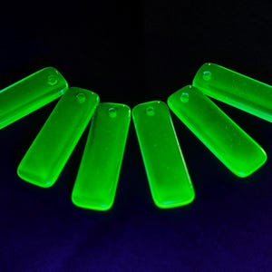 Könnte beinhalten: Sechs neongrüne, rechteckige Glasanhänger mit abgerundeten Kanten sind halbkreisförmig vor einem dunkelblauen Hintergrund angeordnet. Jeder Anhänger hat oben ein kleines Loch zum Auffädeln.