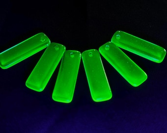 May include: Six luminous green rectangular glass pendants with rounded edges are arranged in a semi-circle against a dark blue background. Each pendant has a small hole at the top for stringing.