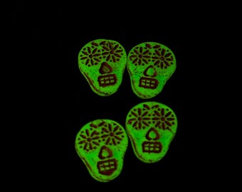 May include: Four skull-shaped beads glowing in a vibrant neon green colour against a dark background. Each bead features intricate dark brown etched details, including floral eye sockets, a triangular nose, and a rectangular grid-patterned mouth.