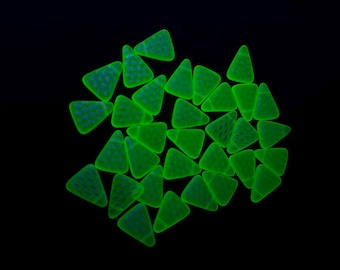 May include: A collection of triangular candy pieces glowing in a vibrant neon green colour against a solid black background. Each piece features a subtle, darker green dotted pattern across its surface, arranged in a scattered, overlapping cluster.