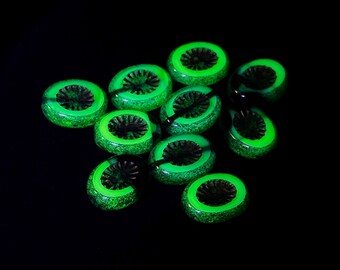 May include: A collection of ten round, flat beads featuring a vibrant neon green speckled outer ring and a dark, textured, star-shaped centre. The beads exhibit a luminous quality against a solid black background, highlighting their circular shape and detailed, radial patterns.