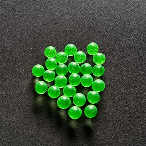 May include: A scattering of translucent green glass marbles on a dark surface. The marbles are spherical and have a slight internal texture, with a bright, vibrant green colour. The background is a textured, dark grey.