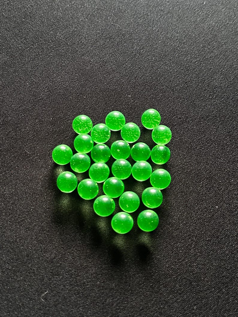 May include: A scattering of translucent green glass marbles on a dark surface. The marbles are spherical and have a slight internal texture, with a bright, vibrant green colour. The background is a textured, dark grey.
