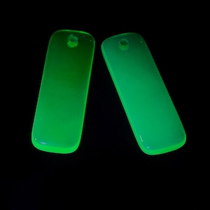 May include: Two rectangular, translucent neon green pendants with rounded corners and a single circular hole at the top of each piece. The objects emit a bright, glowing light against a solid black background, highlighting their smooth, flat surface.