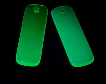May include: Two rectangular, translucent neon green pendants with rounded corners and a single circular hole at the top of each piece. The objects emit a bright, glowing light against a solid black background, highlighting their smooth, flat surface.