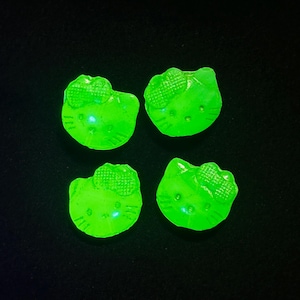 May include: Four small, bright neon green decorative charms shaped like the head of a cat character with a bow on one side. Each charm features embossed facial details including eyes, a nose, and whiskers, glowing intensely against a dark background.
