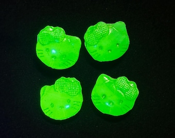 May include: Four small, bright neon green decorative charms shaped like the head of a cat character with a bow on one side. Each charm features embossed facial details including eyes, a nose, and whiskers, glowing intensely against a dark background.