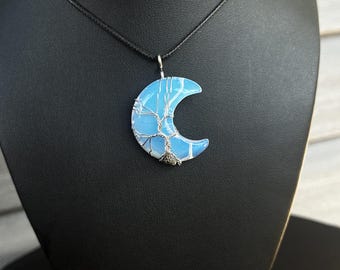 May include: A crescent moon-shaped pendant made of a translucent, milky blue stone wrapped in silver-coloured wire that forms a tree design. The pendant hangs from a thin black cord, showcasing the intricate wire branches and roots.