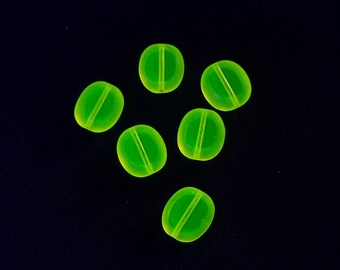 May include: Six luminous green oval beads with a central groove are displayed against a dark backdrop. The translucent beads have a bright, glowing appearance, possibly made of glass or a similar material. These beads are likely for jewellery making.