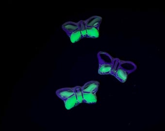 May include: Three small butterfly-shaped charms featuring a translucent purple outer border and a vibrant, glowing neon green centre. These decorative pieces are arranged in a scattered formation against a solid, pitch-black background.