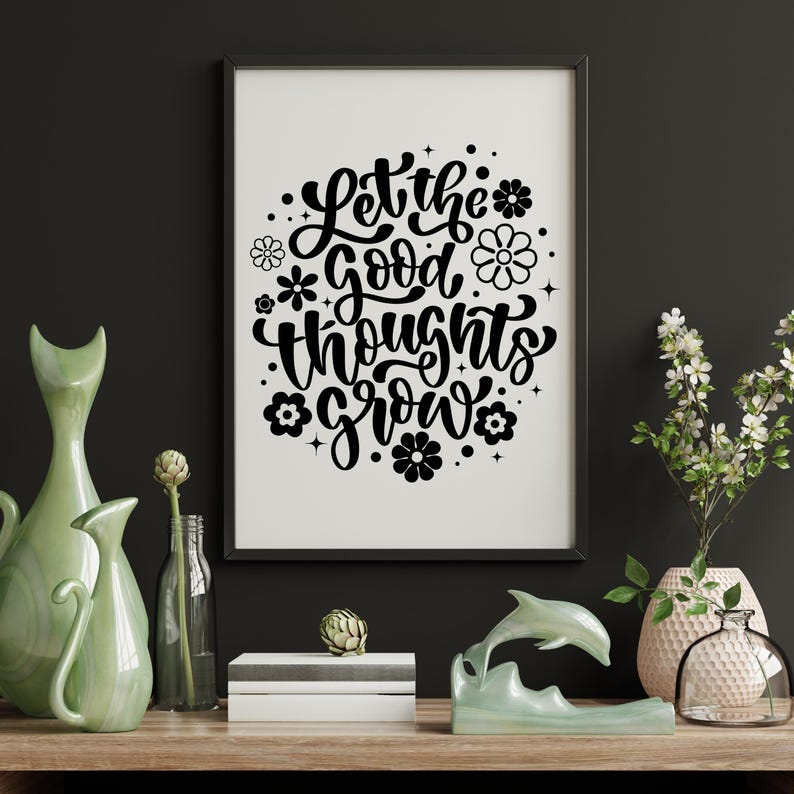 May include: A framed print with the text "Let the good thoughts grow" in black script, surrounded by floral designs. The print is displayed on a wooden surface with green vases, a dolphin sculpture, and a vase of flowers.