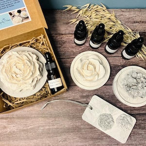 May include: A set of three white ceramic flower diffusers with a small bottle of essential oil. The diffusers are in the shape of roses and peonies. The set is packaged in a brown box with a white label that says "Uniquely Ours".