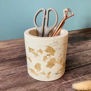 Könnte beinhalten: Weißer Marmor-Bleistifthalter mit goldenen Flocken, mit einer silbernen Schere und einem roségoldenen Stift mit einer diamantförmigen Spitze.
