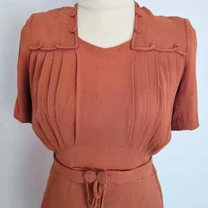 Peut inclure: Robe vintage de couleur rouille avec des manches courtes et une encolure carrée. Le corsage présente des plis verticaux et des détails décoratifs aux épaules et à la taille. La robe comprend une ceinture assortie avec un ornement noué unique.