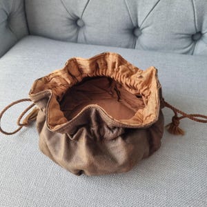 Puede incluir: Un monedero de cuero marrón con un interior marrón claro. El monedero está abierto, revelando el interior, y tiene un cierre de cordón con borlas. El cuero tiene una apariencia texturizada.