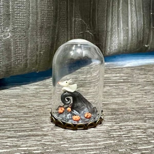 May include: A glass dome containing a black and white figurine of Jack Skellington from The Nightmare Before Christmas. The figurine is perched on a black hill with orange pumpkins.