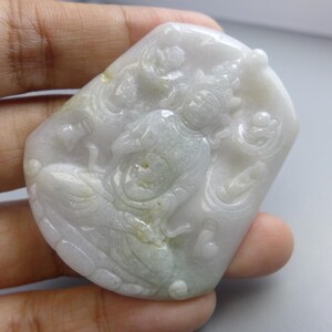 Może przedstawiać: Rzeźbiony wisiorek z jadeitu przedstawiający siedzącą postać z płynącymi szatami i skomplikowanymi detalami. Wisiorek jest głównie biały z subtelnymi zielonymi odcieniami, a rzeźba jest osadzona na gładkiej, wypolerowanej powierzchni. Ogólny projekt jest tradycyjny i ozdobny.