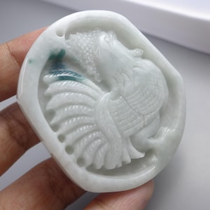 May include: A carved jade pendant featuring a detailed relief of a bird, possibly a phoenix or rooster. The pendant is primarily white with subtle green and blue inclusions. The bird's feathers and form are intricately carved.