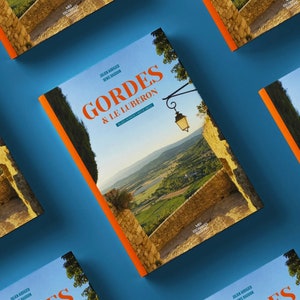 May include: A book cover with an orange border and the title "Gordes & Le Luberon" in large, bold, white letters. The background image is a scenic view of a village in the French countryside.