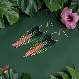 Op de afbeelding: Oorbellen met kralen, goudkleurige ringen en oorhaken. De oorbellen hebben een verloop van groene en roze kralen, met goudkleurige accenten. Ze zijn geplaatst tegen een donkergroene achtergrond met tropische bladeren en een roze bloem.