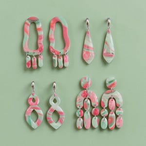 Könnte beinhalten: Sechs Paar bunte Ohrringe mit einem marmorierten Design in Rosa, Grün und Weiß. Die Ohrringe haben verschiedene Formen, darunter Tropfen, offene Ovale und geschichtete Designs, alle mit silberfarbenen Verschlüssen. Präsentiert auf einem hellgrünen Hintergrund.