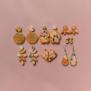 Könnte beinhalten: Eine Auswahl an floralen Ohrringen in verschiedenen Formen, darunter Ohrstecker und Hängeohrringe. Die Ohrringe zeigen orange, rosa und weiße Blumenmuster. Einige haben goldfarbene Details. Der Hintergrund ist rosa.