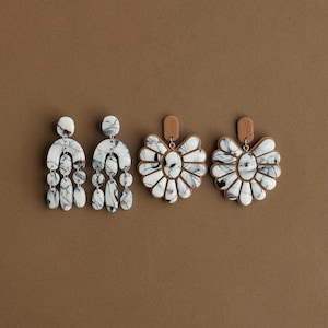 Könnte beinhalten: Drei Paar Ohrringe auf braunem Hintergrund. Zwei Paare sind weiß mit schwarzer Marmorierung, eines mit einem hängenden Design und das andere mit einer floralen Form. Das dritte Paar hat eine florale Form mit braunem Rücken.
