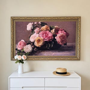 May include: A rectangular wall art print in an ornate gold frame featuring a still life of pink, peach, and cream-colored roses in a dark bowl. Below the frame, a white vase holds three artificial roses, and a straw hat with a black band sits.