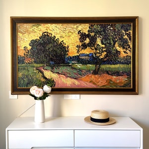 May include: A framed oil painting depicting a landscape with two trees and a house in the distance. The painting is set against a yellow sky with a hint of orange and blue. The trees are green and brown, and the house is white with a brown roof. The painting is framed in a dark brown wood frame.