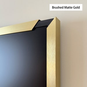 Puede incluir: Primer plano de un marco de imagen de oro mate cepillado con un passe-partout negro. El marco tiene un acabado texturizado sutil.