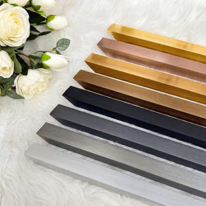 Puede incluir: Una selección de piezas de marcos de fotos de madera en varios colores metálicos y neutros, incluyendo oro, bronce, negro y plata. Los marcos están dispuestos diagonalmente sobre una superficie blanca y esponjosa, con rosas blancas en la esquina superior izquierda.