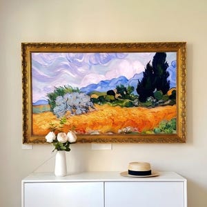 May include: A landscape painting in a gold ornate frame featuring a golden wheat field, a silver-toned tree, and dark green cypress trees under a swirling lavender and white sky. Below sits a white vase with three pale pink roses and a straw hat.