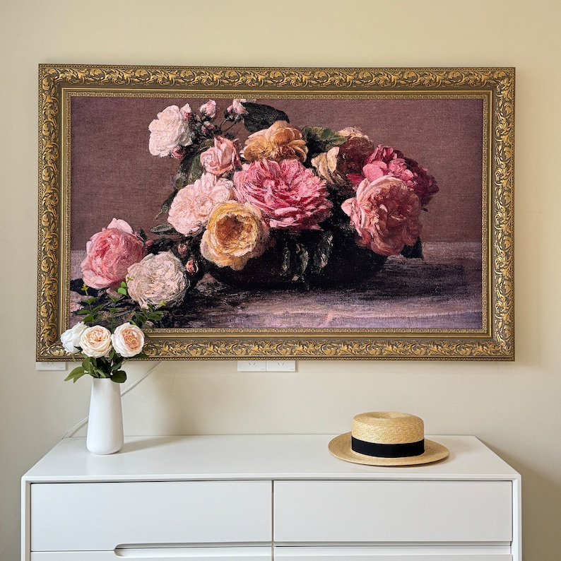 May include: A framed tapestry depicting a bouquet of pink and white roses in a vase. The roses are arranged in a cluster, with some petals overlapping. The background is a dark brown color, and the frame is a gold color.
