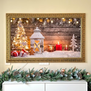 May include: A framed Christmas scene with a rustic wooden background. It features a white lantern, lit candles, a gingerbread man, and a small Christmas tree. Presents and decorative lights add to the festive atmosphere. The frame is gold.