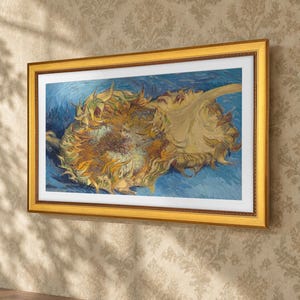 以下が含まれることがあります： フィンセント・ファン・ゴッホのひまわりの絵画の額入りプリント。鮮やかな青い背景に、質感のある黄色と茶色の大きなひまわりの花が2つ描かれており、白いマットボーダーが付いた装飾的なゴールドトーンのフレームに収められています。