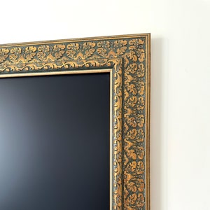 May include: Close-up of a decorative rectangular frame with a dark interior. The frame features an ornate gold and dark green floral design. The frame is against a white wall.