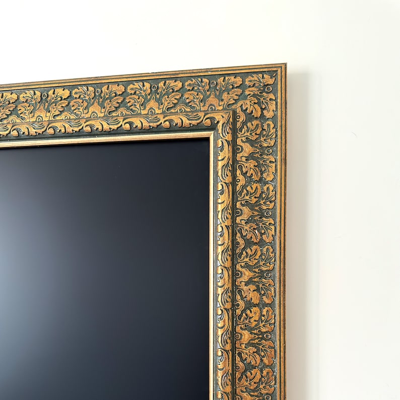 May include: Close-up of a decorative rectangular frame with a dark interior. The frame features an ornate gold and dark green floral design. The frame is against a white wall.
