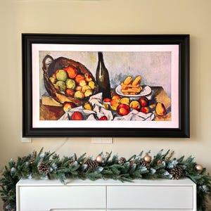 May include: A framed painting of a still life with a basket of apples, a bottle of wine, and a plate of pastries. The painting is in a dark wood frame and is hanging on a wall.