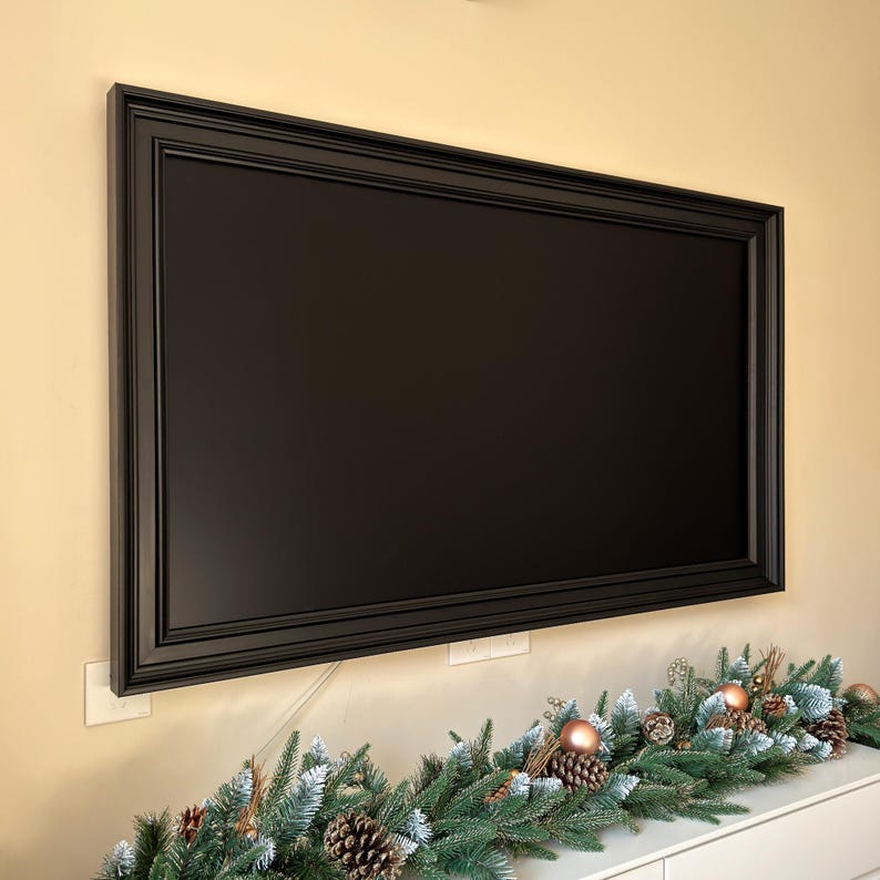 May include: A black framed television mounted on a wall above a white surface with a green garland decorated with pine cones and brown ornaments.