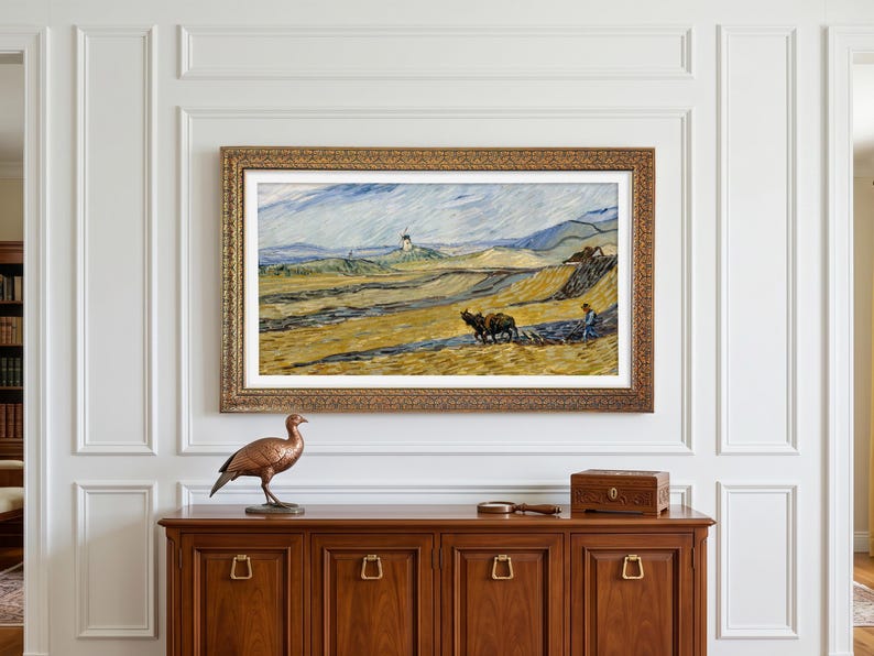 May include: A framed landscape painting with a gold ornate frame hangs above a wooden cabinet. A bronze bird statue, a small wooden box, and a magnifying glass are on the cabinet. The painting depicts a field with a farmer and oxen.
