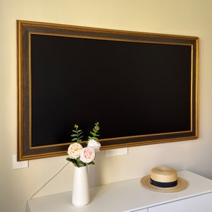 May include: A black framed mirror with a gold trim hangs on a white wall. A vase with pink and white flowers sits on a white table in front of the mirror. A straw hat sits on the table next to the vase.