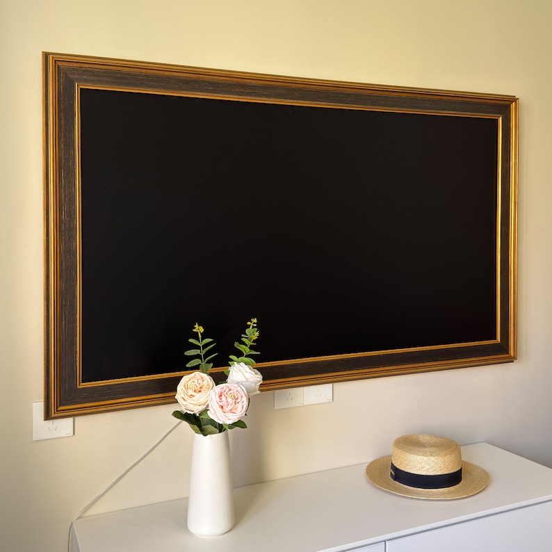 May include: A black framed mirror with a gold trim hangs on a white wall. A vase with pink and white flowers sits on a white table in front of the mirror. A straw hat sits on the table next to the vase.