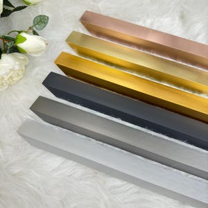 Puede incluir: Cinco soportes de estantería de metal de diferentes colores. Los soportes están dispuestos en una pila de arriba a abajo, con el soporte superior en oro rosa, seguido de oro, latón, gris oscuro y gris claro.