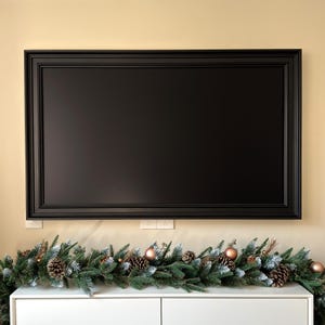 May include: A black framed television mounted on a wall above a white shelf with a green garland decorated with pine cones and gold ornaments.