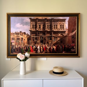 May include: A framed painting depicting a large crowd of people gathered in a public square. The scene is set in a European city with a large building in the background. The painting is in a gold frame and is hanging on a white wall.