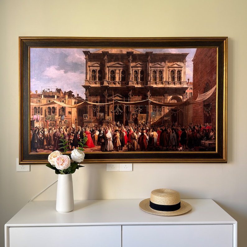 May include: A framed painting depicting a large crowd of people gathered in a public square. The scene is set in a European city with a large building in the background. The painting is in a gold frame and is hanging on a white wall.
