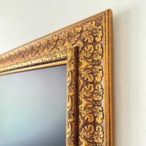 May include: Close-up of a rectangular gold-colored picture frame with intricate leaf and scroll carvings. The frame has a dark brown inner edge and a light gold outer edge, creating a contrast. The frame is mounted on a white wall.
