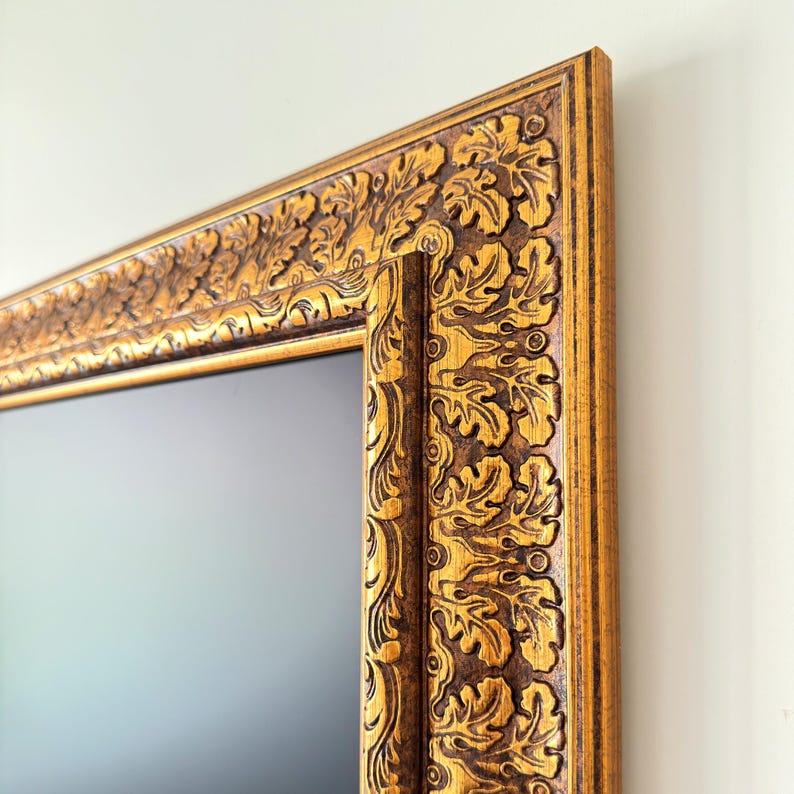 May include: Close-up of a rectangular gold-colored picture frame with intricate leaf and scroll carvings. The frame has a dark brown inner edge and a light gold outer edge, creating a contrast. The frame is mounted on a white wall.